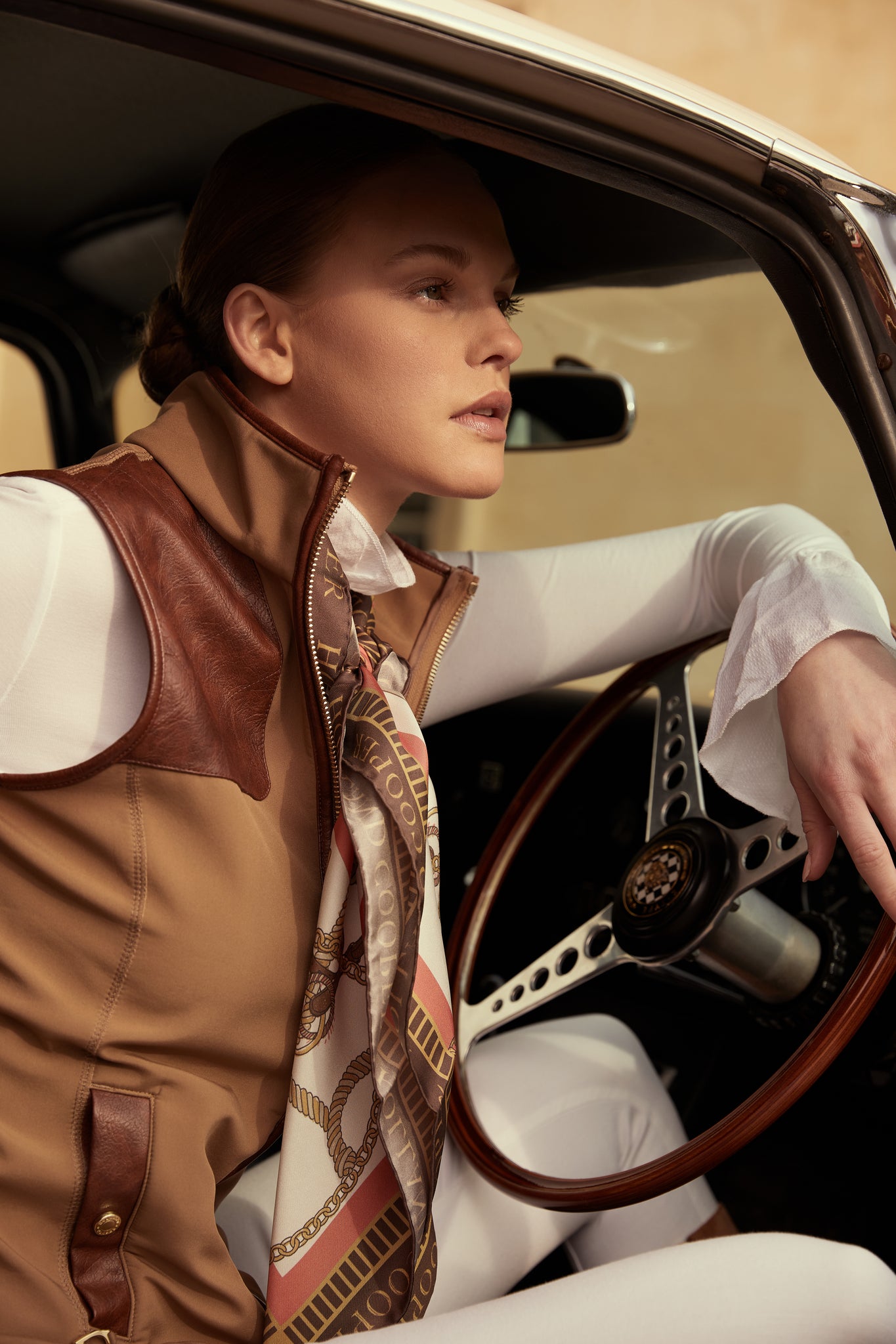 womens light brown gilet with dark brown leather seams along the arm holes pockets and down the zip with a gun patch on the shoulder and an embroidered logo worn with a scarf a white shirt and white skinny jeans