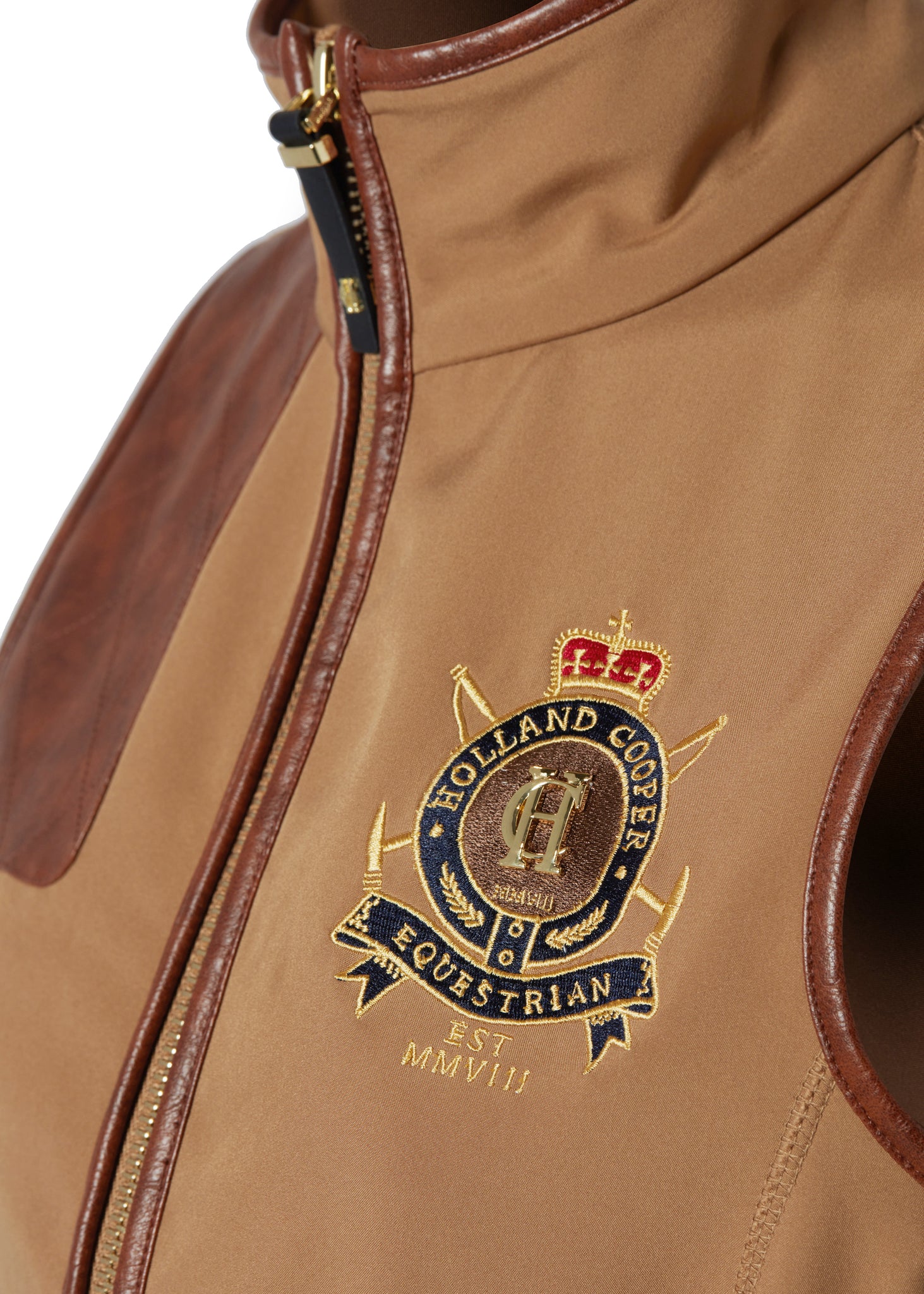 detail shot of embroidered logo on the womens light brown gilet with dark brown leather seams along the arm holes pockets and down the zip with a gun patch on the shoulder